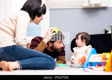 Homme et femme heureux dans des vêtements décontractés assis sur le sol et jouant avec la petite fille tout en passant du temps à la maison ensemble Banque D'Images