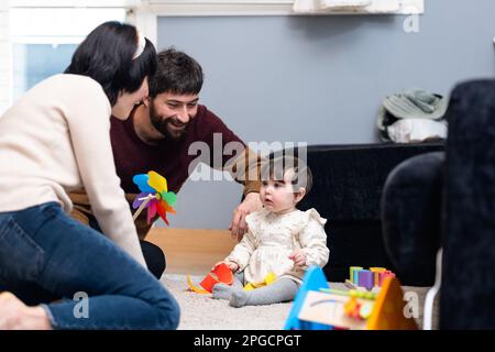 Homme et femme heureux dans des vêtements décontractés assis sur le sol et jouant avec la petite fille tout en passant du temps à la maison ensemble Banque D'Images