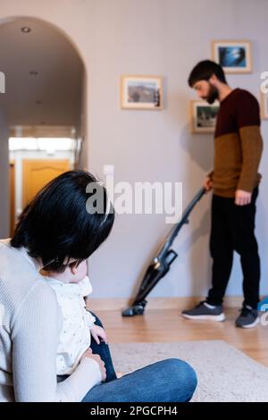 Mère méconnue assise avec une petite fille et un père qui nettoie le sol avec un aspirateur en arrière-plan à la maison Banque D'Images