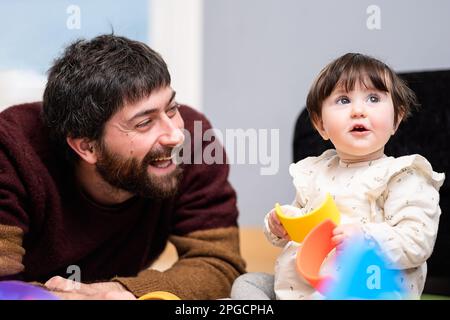 Homme heureux dans des vêtements décontractés assis sur le sol et jouant avec la petite fille tout en passant du temps à la maison ensemble Banque D'Images