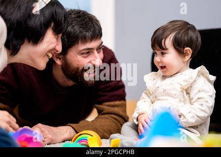 Homme et femme heureux dans des vêtements décontractés assis sur le sol et jouant avec la petite fille tout en passant du temps à la maison ensemble Banque D'Images