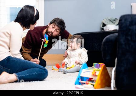 Homme et femme heureux dans des vêtements décontractés assis sur le sol et jouant avec la petite fille tout en passant du temps à la maison ensemble Banque D'Images