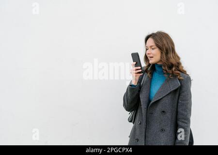Jeune femme en manteau enregistrant un message vocal sur un smartphone debout regardant l'écran sur fond blanc Banque D'Images