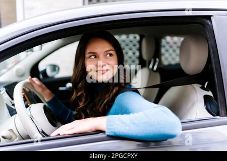 Vue latérale d'une jeune femme souriante dans une voiture à col roulé avec intérieur blanc tout en regardant loin Banque D'Images