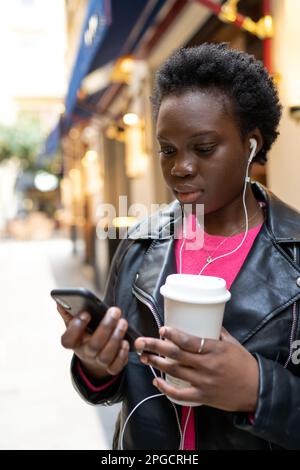 Femme afro-américaine naviguant sur un smartphone tout en se tenant debout et en écoutant de la musique avec des écouteurs dans la rue Banque D'Images