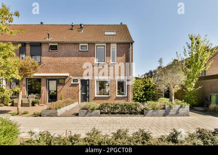 Design extérieur de la maison résidentielle en brique avec fenêtres et portes situées dans le quartier de banlieue avec des arbres verts et des plantes sous ciel bleu sans nuages Banque D'Images