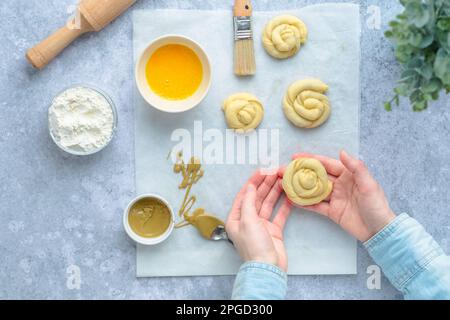 Main de la femme tenant un petit pain en spirale avant de cuire. Biscuits suédois, œufs battus, farine, roulailler une cuillère avec de la crème pistache sur papier parchemin Banque D'Images