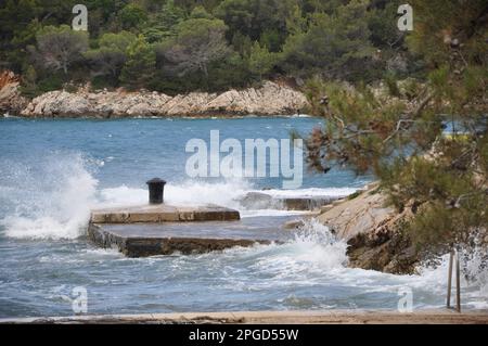Jetée en pierre avec un bollard rouillé. Rolard d'amarrage en fer délabré complètement rouillé au milieu de la jetée en pierre. Pins sur la côte. Banque D'Images