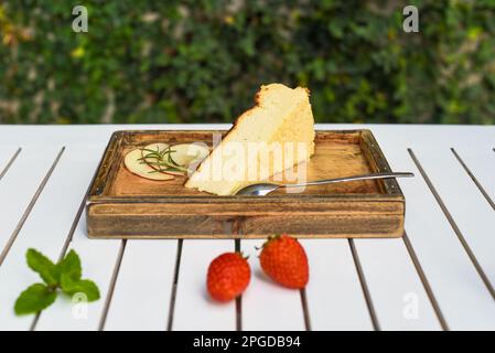 Morceau de cheesecake sur une planche en bois avec fraise et menthe sur table en bois blanc avec feuilles vertes et espace de copie Banque D'Images