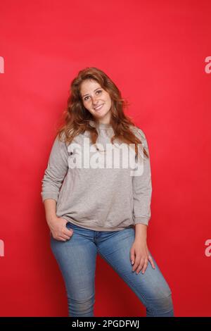 Femme brune mûre attirante en chandail gris et jeans bleus debout sur fond rouge de mur de studio Banque D'Images