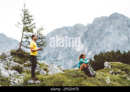 Couple de randonneurs prenant un repos dans les montagnes, femme assis sur l'herbe et homme regardant vers le haut dans le ciel Banque D'Images