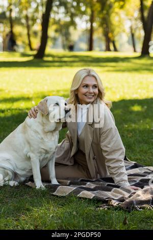 bonne femme d'âge moyen avec des cheveux blonds qui câblent le chien labrador tout en étant assis sur une couverture dans le parc, image de stock Banque D'Images