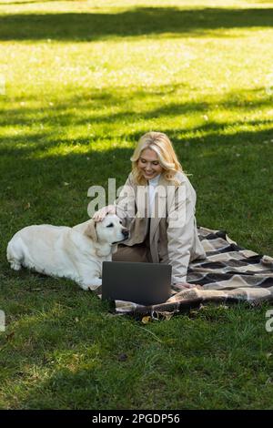 bonne femme d'âge moyen qui pète un chien du labrador tout en regardant un film sur un ordinateur portable et en étant assise sur une couverture dans le parc, image de stock Banque D'Images