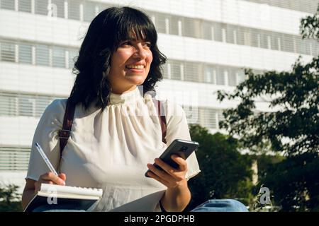 jeune étudiante latine d'origine vénézuélienne, cheveux courts et vêtements décontractés, souriante et réfléchie assis à l'extérieur de l'université avec son phon Banque D'Images