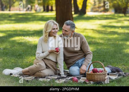 gaie femme d'âge moyen tenant une pomme mûre près de mari barbu pendant le pique-nique dans le parc, image de stock Banque D'Images