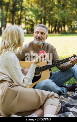 joyeux homme d'âge moyen avec une barbe grise jouant de la guitare acoustique près de la femme blonde avec pomme rouge pendant le pique-nique dans le parc, image de stock Banque D'Images