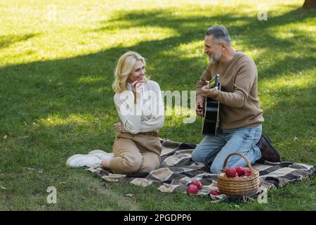 joyeux homme d'âge moyen avec barbe grise jouant de la guitare acoustique près de femme insouciante avec pomme pendant le pique-nique dans le parc, image de stock Banque D'Images