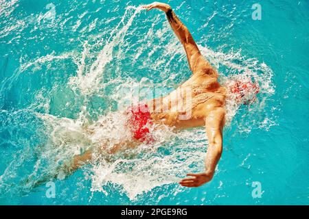 Un homme nage dans la piscine dans une casquette rouge et des lunettes parmi les rayons du soleil, train technique de natation, vue de dessus, style papillon, espace d'imitation Banque D'Images