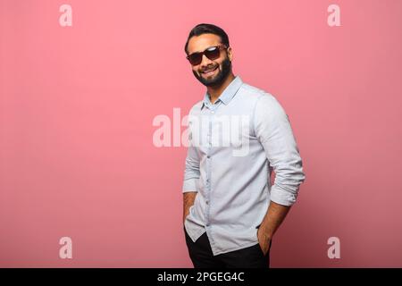 Joli homme indien barbu portant des lunettes de soleil dans un Jean décontracté chemise regardant l'appareil photo et souriant isolé sur le rose. Un homme charismatique du Moyen-Orient se tient dans les poches Banque D'Images