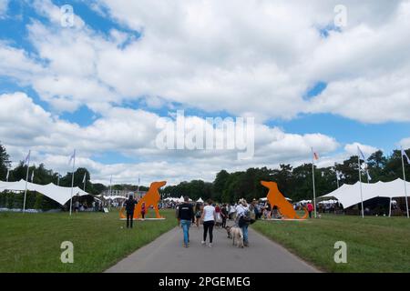 Les gens qui marchent devant deux énormes silhouettes en bois orange de Hounds à l'entrée de Goodwoof, les Kennels, Goodwood, West Sussex, Royaume-Uni Banque D'Images