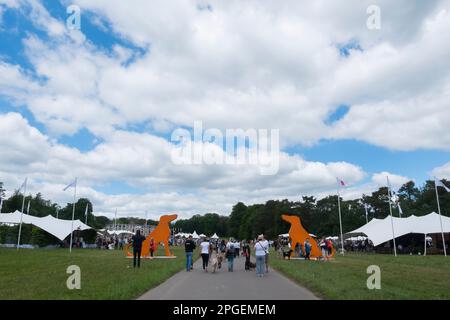 Les gens qui marchent devant deux énormes silhouettes en bois orange de Hounds à l'entrée de Goodwoof, les Kennels, Goodwood, West Sussex, Royaume-Uni Banque D'Images