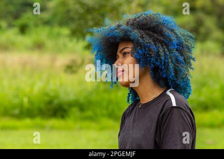 Goiania, Goias, Brésil – 20 mars 2023: Photo d'une jeune femme noire aux cheveux afro, bleu teint, avec un petit sourire sur son visage, en profil avec flou Banque D'Images