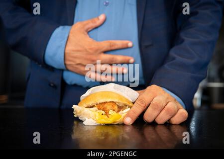 Concept de manger des aliments malsains. homme tenant un hamburger gras et touchant son estomac. Gastrite ou ulcère chronique. Indigestion des intestins de Banque D'Images