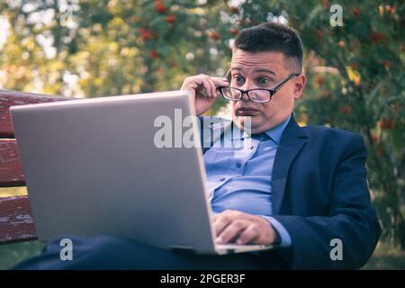 Le jeune homme a surpris avec son ordinateur portable. l'homme d'affaires se repose avec un ordinateur portable dans le parc en été regarder la vidéo Banque D'Images