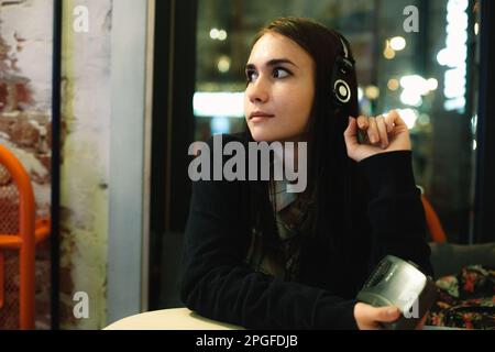 Jeune femme écoutant de la musique dans un casque assis dans un café la nuit Banque D'Images