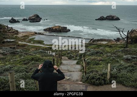 Un homme se promenant le long des chemins restaurés des ruines des bains de Sutro, avec l'océan Pacifique qui s'étend derrière eux et des mouettes qui s'envolent au-dessus. Les thermes Sutro, un site historique qui abritait autrefois le plus grand complexe de piscines intérieures du monde, continuent de captiver les habitants et les touristes avec leur histoire fascinante et leurs vues imprenables sur l'océan. Construit en 1896 par l'ancien maire de San Francisco Adolph Sutro, les bains étaient une grande attraction qui a accueilli des visiteurs du monde entier. Le complexe était une merveille d'ingénierie, avec six piscines d'eau salée, un immense dôme en verre et des sièges Banque D'Images