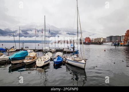 Scène de la nature de la buée et voilier abrité dans le petit port et la jetée en eau d'hiver et les mélèzes d'orange à distance et la ville de Locarno Banque D'Images