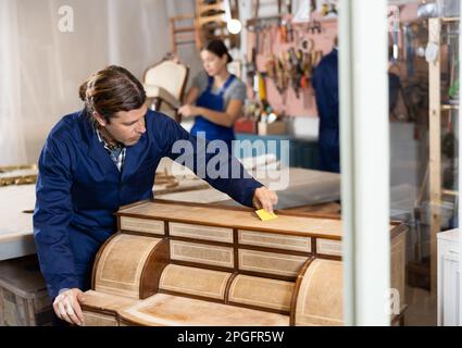 Un homme restaurateur expérimenté âgé restaure soigneusement et soigneusement la poitrine des tiroirs dans l'atelier de restauration Banque D'Images