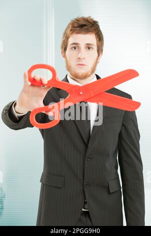 Un travailleur de bureau à fines rayures noires avec une barbe et de courts cheveux rouges tient un gros ciseau rouge. Concept de coupe. Réduction du personnel, des coûts et des dépenses Banque D'Images