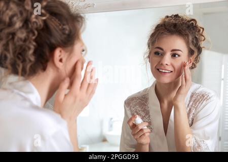 Belle femme appliquant de la crème hydratante près du miroir dans la salle de bains Banque D'Images