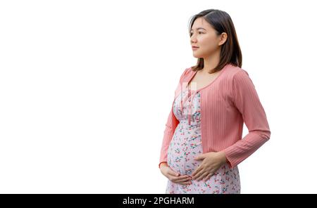 Une jeune femme asiatique heureuse, vêtue de vêtements de maternité, souriant et debout devant un fond blanc tenant son ventre tout en pensant à son bébé Banque D'Images