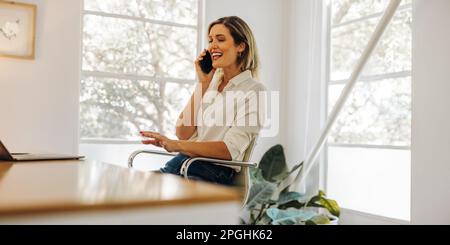 Femme d'affaires souriante prenant un appel téléphonique dans son bureau à domicile. Bonne jeune femme d'affaires communiquant avec ses partenaires d'affaires tout en travaillant à distance Banque D'Images