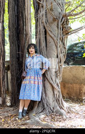 Une fille souriante brune de vingt-cinq ans vêtue d'une robe à motifs bleu vintage avec un petit sac à main appuyé contre le tronc d'une grande fiche Banque D'Images