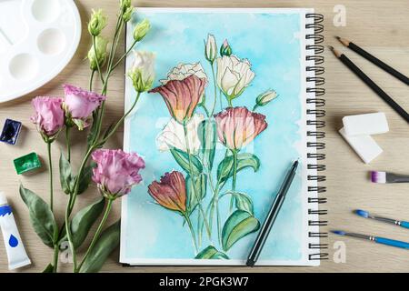 Peinture d'eustomas dans le carnet d'esquisses, fleurs et fournitures d'art sur table en bois, pose plate Banque D'Images