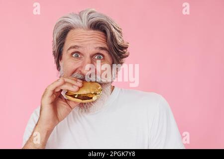 Mange un délicieux hamburger. Homme moderne et élégant avec cheveux gris et barbe à l'intérieur Banque D'Images