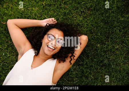 Vue de dessus d'une femme africaine joyeuse en lunettes écoutant de la musique avec des écouteurs tout en se reposant sur l'herbe dans le parc Banque D'Images