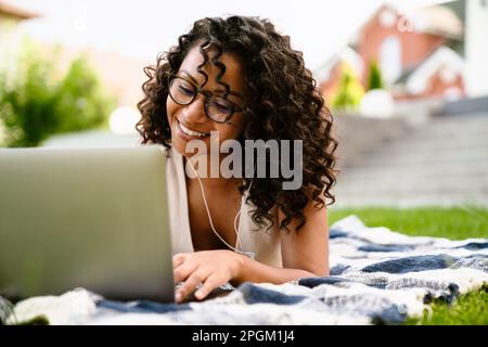 Joyeuse jeune afro-américaine femme dans des écouteurs en utilisant un ordinateur portable tout en étant allongé sur une couverture sur l'herbe verte Banque D'Images