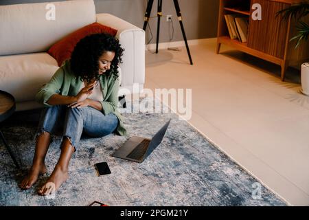 Jeune femme africaine joyeuse ayant le chat vidéo à l'aide d'un ordinateur portable tout en étant assise sur le sol à la maison Banque D'Images