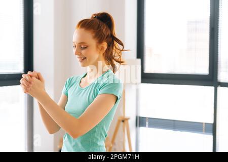 Vue latérale d'une jeune femme sportive heureuse faisant un effort poignet avant l'entraînement de remise en forme au bureau à la maison près de la fenêtre. Femelle à tête rouge positive Banque D'Images