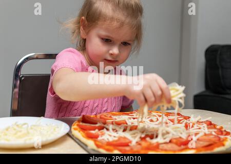 Repas de cuisine pour petit enfant à la maison assis à la table dans la cuisine à la maison. Petite fille faisant de la pizza, ajoutant du fromage. Banque D'Images