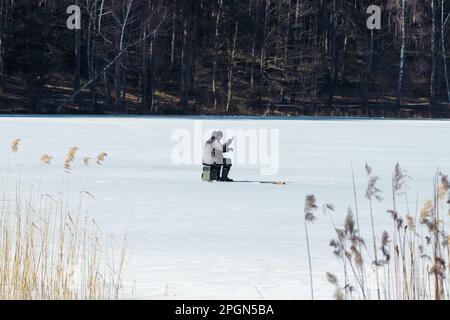 Pêcheur pêchant sur un lac gelé au début du printemps avec un poteau de pêche, une tarière de glace et du matériel de pêche Banque D'Images