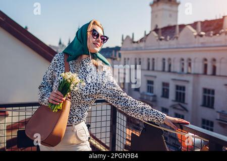 Femme heureuse élégante portant un châle rétro vert avec des lunettes de soleil tenant des fleurs printanières et un sac à main appréciant l'architecture de la ville. Classique vintage extérieur f Banque D'Images