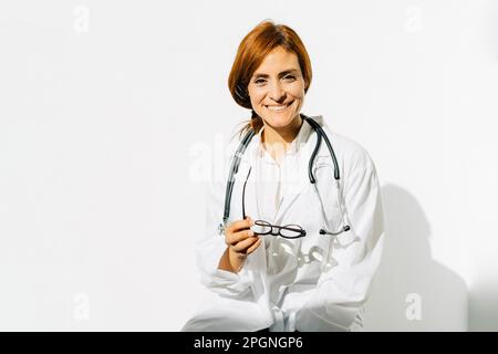 Bonne femme médecin avec des lunettes et un stéthoscope devant le mur blanc Banque D'Images