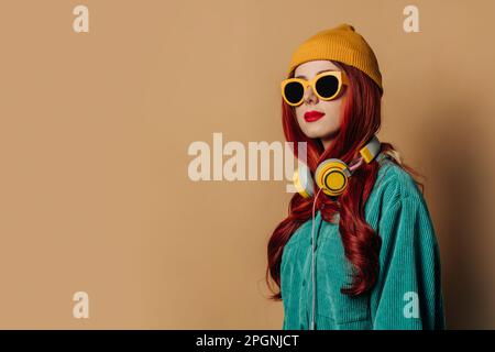 Femme souriante à tête rouge portant un chapeau en tricot et des lunettes de soleil sur fond marron Banque D'Images