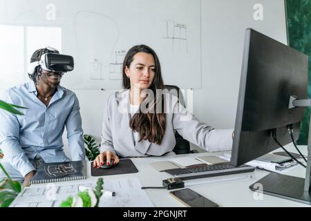 Architecte utilisant un ordinateur avec un collègue portant un casque de réalité virtuelle au bureau Banque D'Images