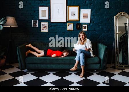 Femme avec des documents assis par un garçon allongé sur un canapé au studio Banque D'Images